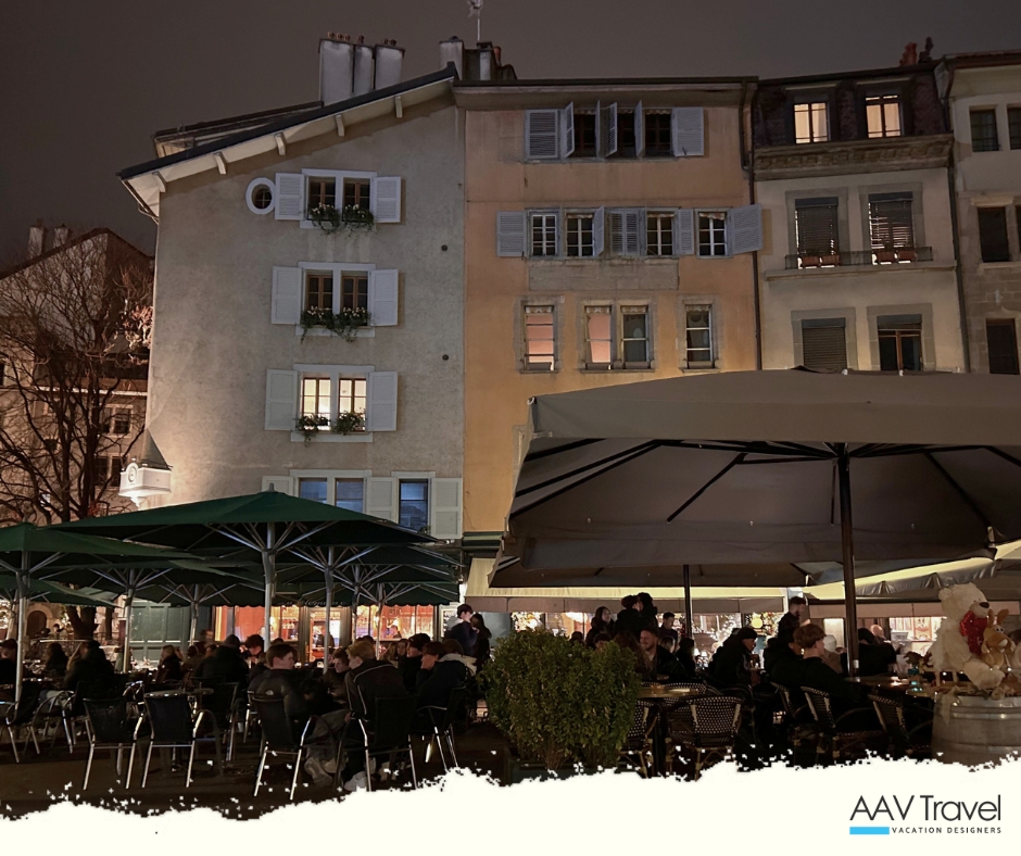 Nighttime scene of a bustling outdoor cafe with patrons seated under large umbrellas, surrounded by historic buildings in a European city.