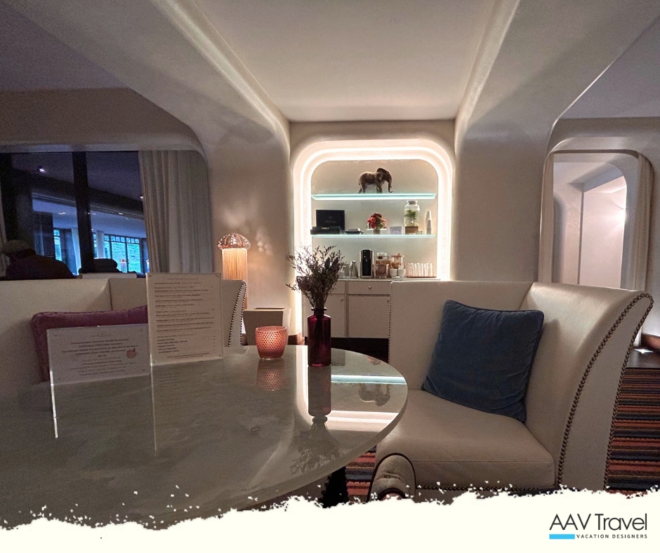 A cozy, elegant interior of a hotel spa, featuring a round table with a textured surface, stylish seating, and soft lighting. A vase of dried flowers and a lit candle add a warm ambiance, while shelves in the background display decorative items.